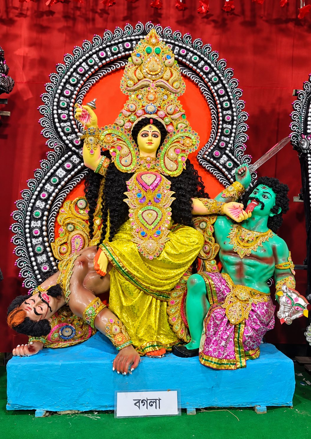 Maa Bagalamukhi (Maa Bogola) at the Chetla Dasamahavidya Puja — the golden-robed Goddess seated, holding the tongue of the restrained opponent, with her nameplate below