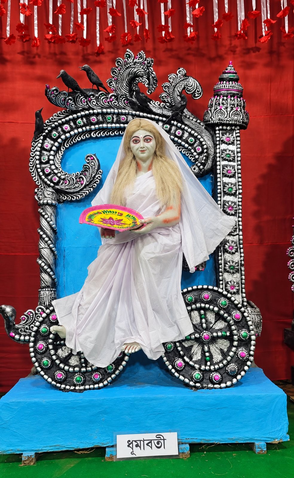 Maa Dhumavati — the pale Goddess in white widow's robes holding the winnowing fan, crows perched on her black-and-silver arch, with her nameplate 'ধূমাবতী' below