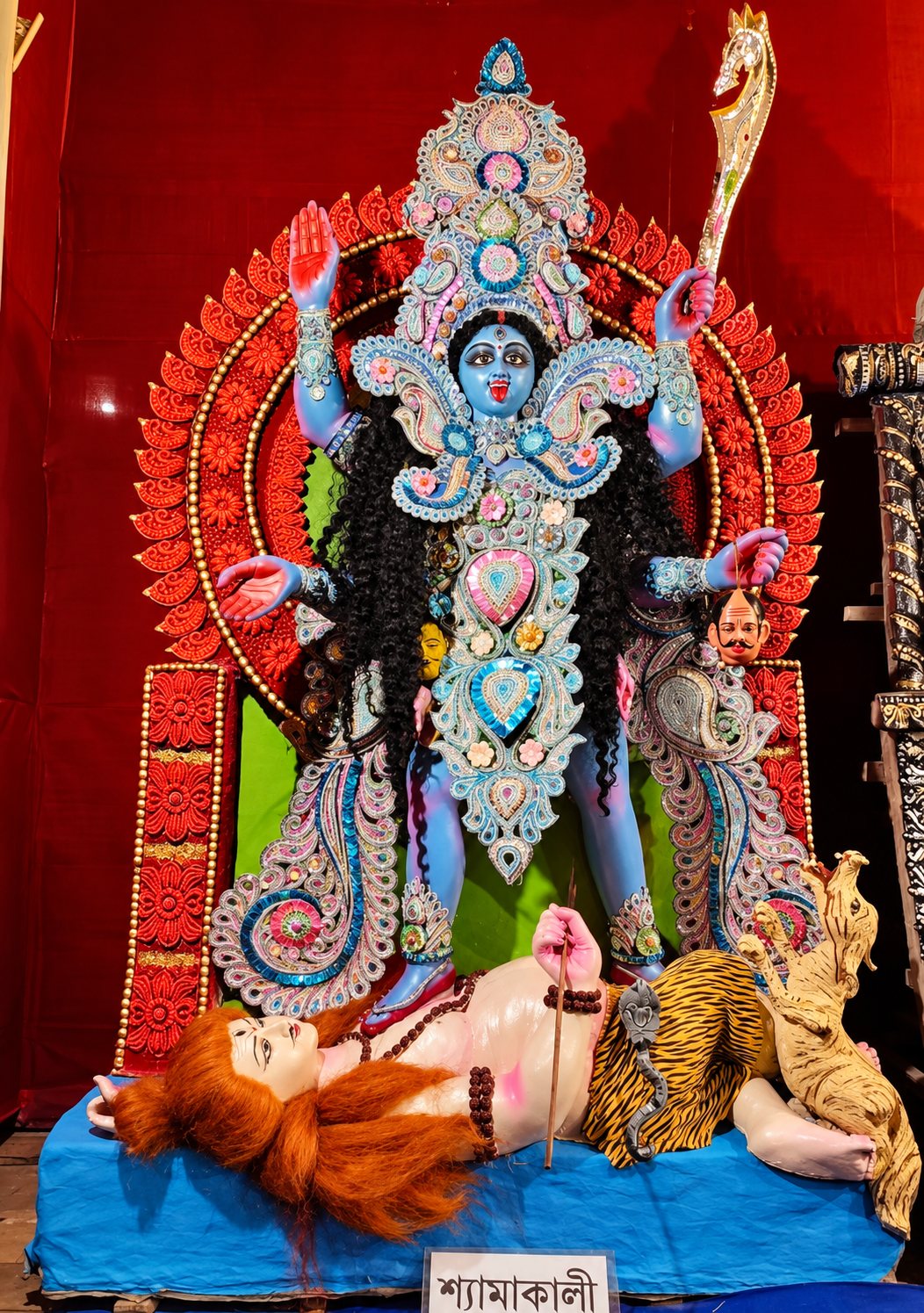 Shyama Maa — the blue four-armed Goddess with sword and blessing hand, standing upon the reclining Shiva, her nameplate 'শ্যামাকালী' below
