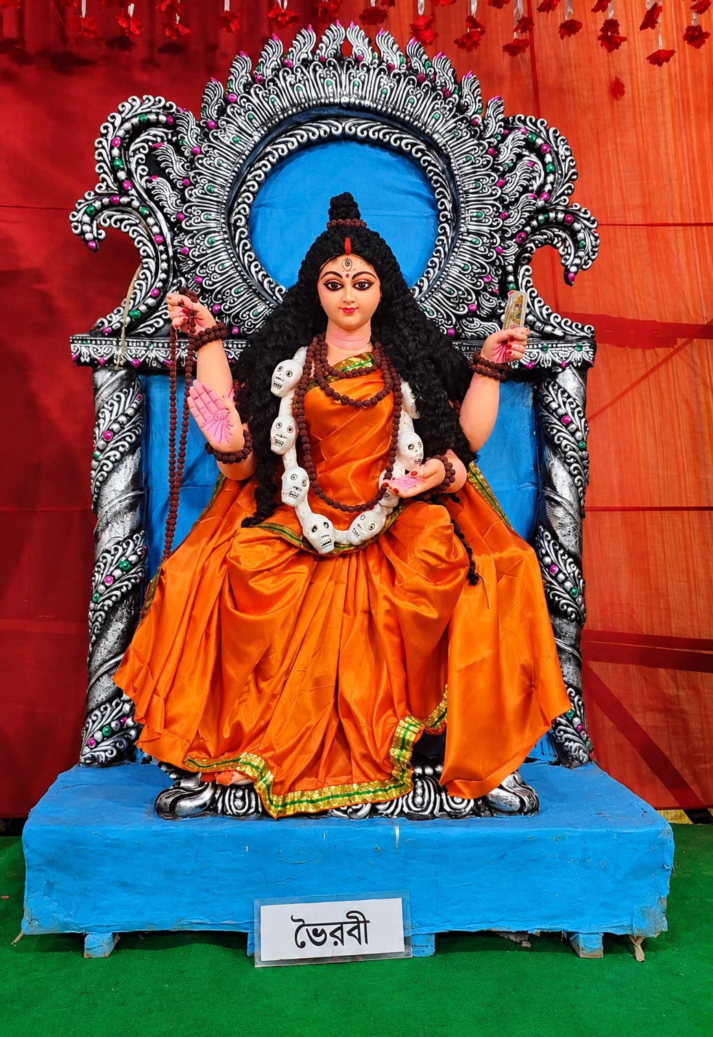 Maa Bhairavi seated in orange robes with rudraksha beads and a garland of skulls, her nameplate 'ভৈরবী' below