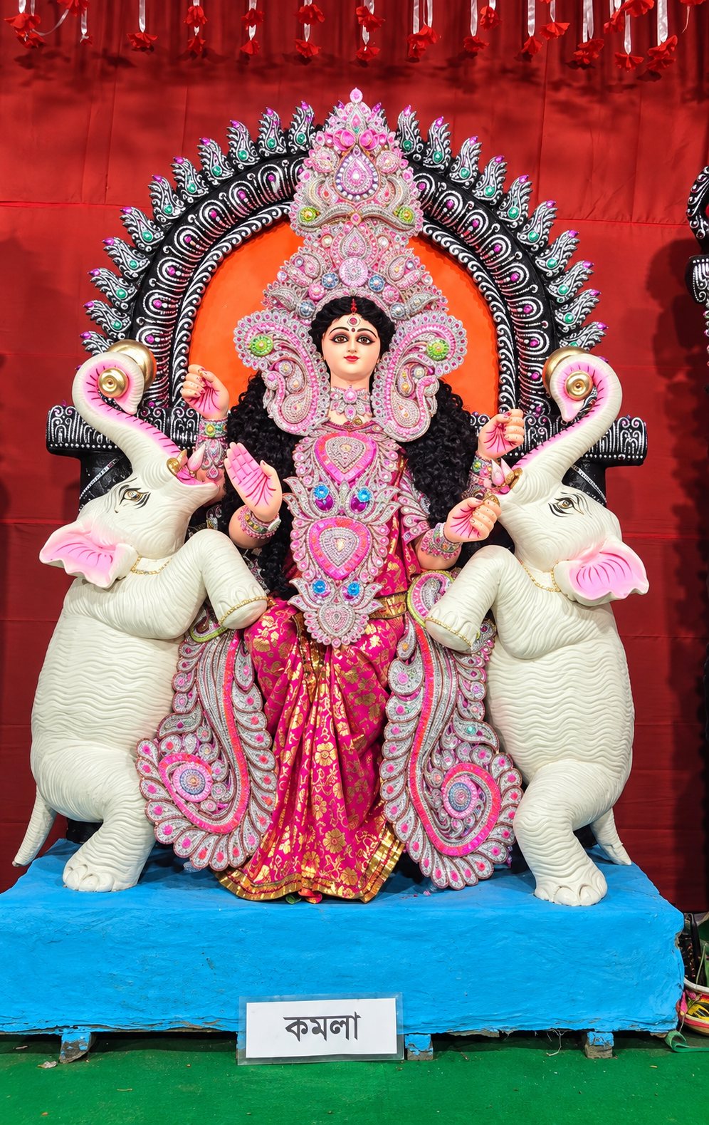 Maa Kamala (Maa Komola) in pink and gold, flanked by two white elephants, with her nameplate 'কমলা' below