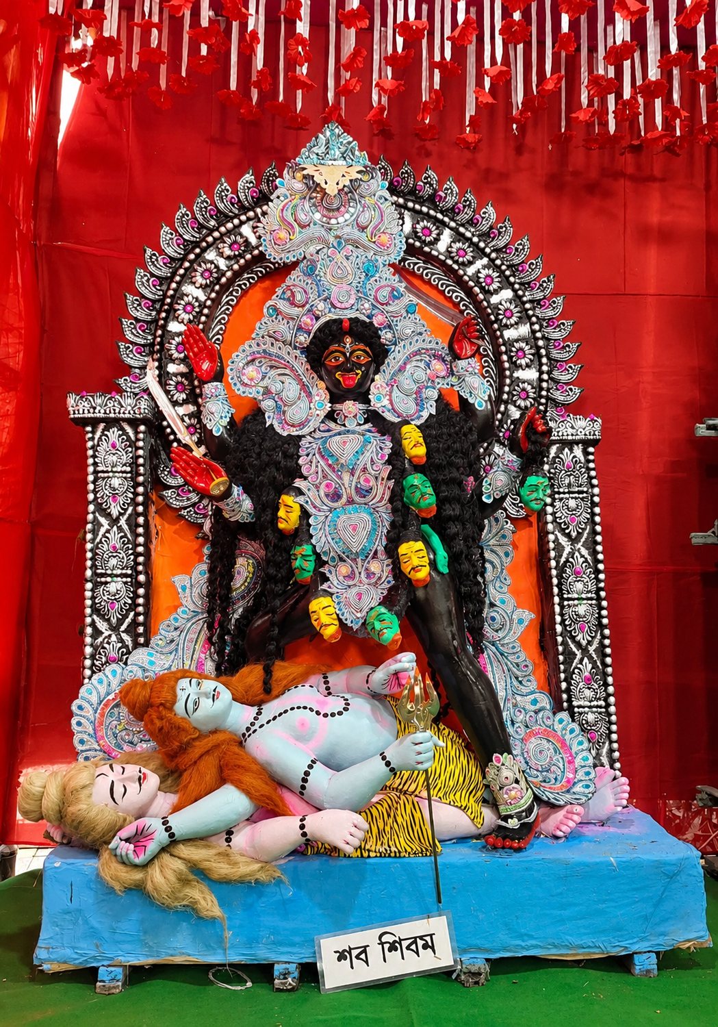 Maa Shabashivam Kali — the first Mahavidya of the Chetla celebration, standing in her fierce form upon the reclining figures, with her nameplate 'শবশিবম' below