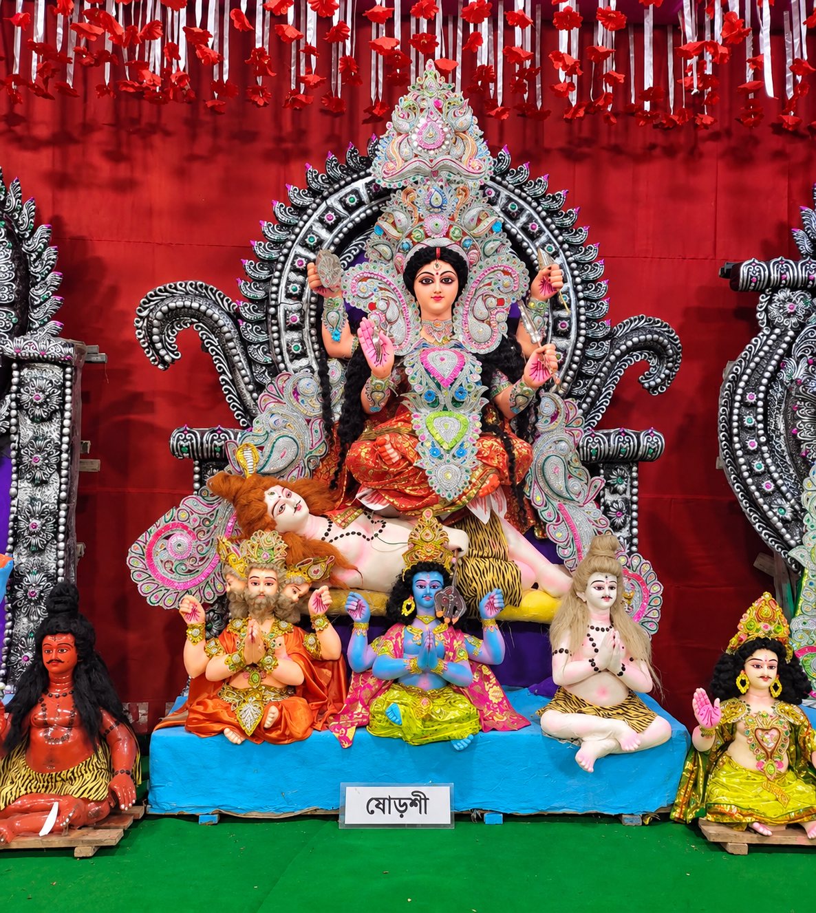 Maa Shoroshi (Shodashi) enthroned in red and silver, with the assembled deities at her feet and her nameplate 'ষোড়শী' below