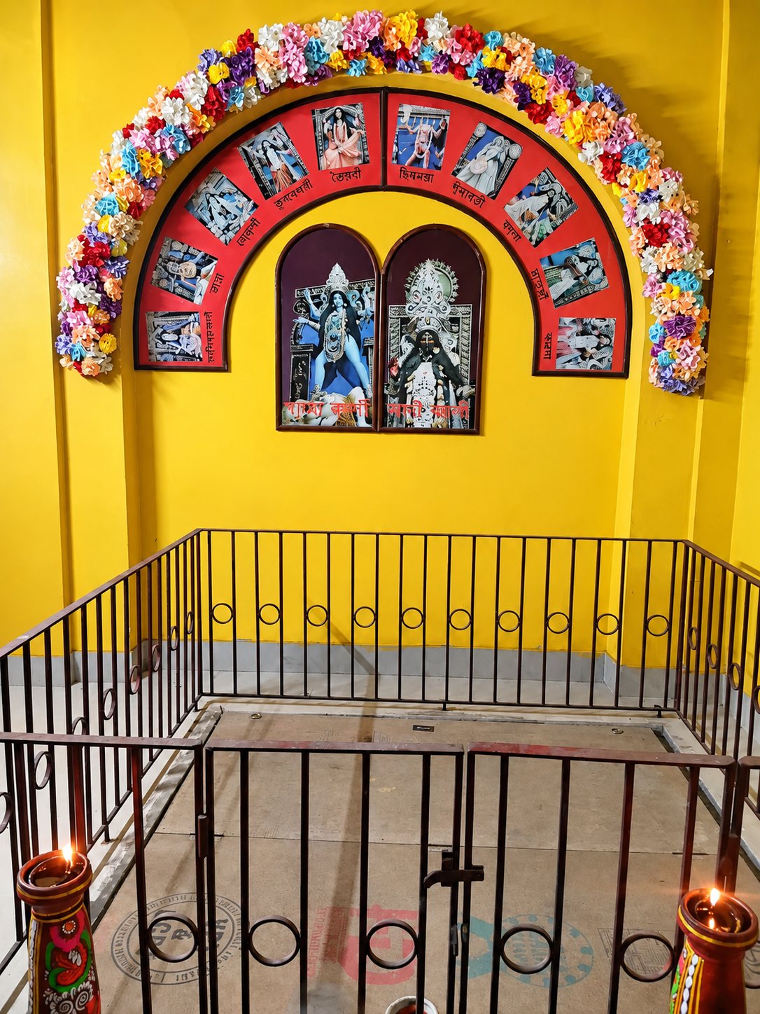 The sanctum at Chetla — the yellow-walled shrine where the Puja takes place, with photographs of the ten Mahavidyas arranged in an arch above the railed platform and lamps burning below